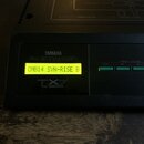 Detailní pohled na ovládací panel černého elektronického zařízení. Na panelu jsou nápisy YAMAHA, FM EXPANDER a TX7 TONE GENERATOR. Rozsvícený displej…