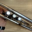 Detailní pohled na tři stříbrné ventily trumpety. Povrch nástroje vykazuje drobné škrábance a známky používání. V pozadí je vidět dřevěná podlaha a č…