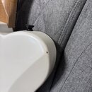 Detail zadní strany bílé elektrické kytary. Na povrchu je vidět malý tmavý odštípnutý kousek laku. V horní části je umístěn černý šroub s plastovou h…