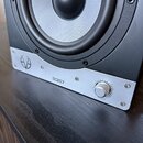 Detailní pohled na spodní část černého studiového monitoru s šedým kovovým předním panelem. Na panelu je vlevo logo ve tvaru stylizovaného kruhu, upr…