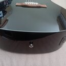 Detail spodní části černé akustické kytary s vysokým leskem. Na boku korpusu je umístěn konektor pro kabel a ovládací panel s nečitelným nápisem. V h…