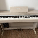 Digitální piano v krémové barvě s povrchem imitujícím světlé dřevo. Nad klávesnicí je umístěn nápis YAMAHA a na hudebním stojanu je vidět vybledlé bí…