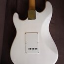 Zadní strana bílé elektrické kytary s tvarem typu Stratocaster. Na těle je vidět kovová krytka krkovnice a bílý plastový kryt elektroniky s řadou šes…