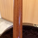 Detailní pohled na hmatník nebo krk strunného nástroje z reddish-brown dřeva s viditelnou sletovanou strukturou. Povrch je lakovaný a lesklý. V pozad…
