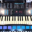 Černý digitální klávesový nástroj s bílými a černými klávesami. Na horním panelu je nápis MUSIC STATION, digitální displej zobrazující červená čísla…