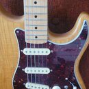 Elektrická kytara s přírodním dřevěným povrchem těla a světlou hmatníkem. Nástroj má tmavě červený pickguard s texturou želvy, tři bílé snímače typu…