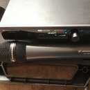 Černý bezdrátový mikrofon s mřížkou a receiver v kovovém šasi. Na obou zařízeních je bílý nápis SENNHEISER a model ewD1 Digital. Displej receiveru zo…
