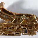Detailní pohled na zlatý saxofon z mosazi s lesklým povrchem. Na korbide je vyryto logo a nápisy "YAMAHA" a "S/N 321...". Nástroj má červené perleťov…