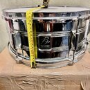 Stříbrný chromovaný buben s bílým koženem, který stojí na kartonovém krabici. Na přední straně je černé logo s nápisem „PD PERFORMANCE PERCUSSION“. K…