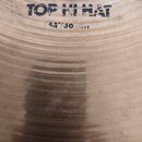 Detail povrchu mosazného činele s viditelnými koncentrickými drážkami. V horní části je modrý nápis TOP HI HAT a pod ním označení 14"/30 ...1. Ve spo…