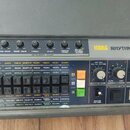 Šedý rytmus stroj s nápisem KORG RHYTHM 55B. Horní panel obsahuje šest otočných regulátorů hlasitosti s popisky Bass Drum, Snare Drum, Cymbal, Hi-Hat…