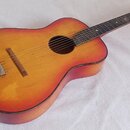 Akustická kytara s žlutě-oranžovým přechodem barvy (sunburst) na deskách a tmavě hnědými boky. Nástroj vykazuje známky opotřebení v podobě škrábanců…