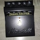 Černý kytarový efekt pedál s nápisy Gain, Bass, Contour, Treble a Volume. Uprostřed je velký bílý nápis SHRED MASTER a ve spodní části vy embossed lo…
