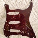 Tmavě červený pickguard s texturou želvy (tortoise shell) určený pro elektrickou kytaru. Má vyřezané otvory pro tři snímače a několik otvorů pro šrou…