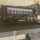 Elektronický klavír v černém tvrdém kufru. Na přístroji je nápis YAMAHA a modelové označení SK10 SYMPHONIC ELECTRONIC PIANO. Přístroj má dřevěné bočn…