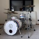 Bílá bicí souprava s modrým a červeným pruhem. Na přední straně basového bubnu je logo s nápisem SINCE 2004, DC DRUMS a QUALITY INSTRUMENTS. Souprava…
