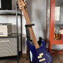 Modrá elektrická basová kytara s bílým pickguardem, umístěná v černém stojanu. Na hlavě hmatníku je vidět nápis "Squier". Tělo nástroje má dva bílé s…