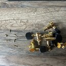 Sada ladíček v zlatém provedení s černými plastovými knoflíky. K ladítkům jsou přiloženy malé zlaté šroubky. Všechny díly leží na dřevěném povrchu.