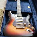 Elektrická kytara typu Stratocaster v barvě sunburst s tmavým hmatníkem a bílým pickguardem. Nástroj je umístěn v otevřeném hnědém kufru s modrým vni…