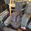 Fotografie zobrazuje hromadu starších černých audio systémů, boomboxů a reproduktorů v různém stavu. Na jednom z přístrojů je viditelný nápis CS-200…