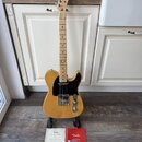 Elektrická kytara s přírodním dřevěným povrchem, černým pickguardem a javorovým hmatníkem. Na hlavě je vidět nápis Fender STRATOCASTER. Kytara stojí…