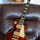 Elektrická kytara typu Les Paul v tmavě červené barvě s bílým pickguardem a krémovými rámečky snímačů. Na hlavě hmatníku je nápis Epiphone a podpis L…