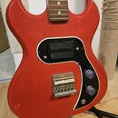 Červená elektrická kytara s dvojsekmovým tělem a hnědým hmatníkem. Nástroj má černý pickguard, dva fialové oválné knofíky a stříbrný most s šestimi s…