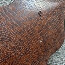 Detailní záběr na hnědý materiál s texturou krokodýlí kůže. Povrch vykazuje známky opotřebení v podobě několika drobných oděraných míst, kde je vidět…