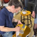 Muž v modrém tričku pracuje na žluté elektrické kytáře s bílým pickguardem a chromovaným hardwarem. Kytara leží v jeho klíně a on do ní zapisuje nebo…