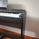 Černý digitální piano s klávesnicí a integrovaným stojanem. Nástroj je umístěn u bílé stěny na oranžovém koberci. Povrch korpusu je matný, bez vidite…