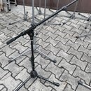 Fotografie zobrazuje několik černých kovových stativů na mikrofony s trojnožkou, umístěných na dlažbové ploše. Na jedné z nožek předního stativu je b…