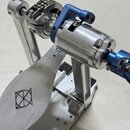 Detailní pohled na kovový mechanismus bubnového pedálu v stříbrné a modré barvě. Na stříbrném šlapadle je vyryto logo znázornující kroužek s přeškrtn…