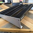 Stříbrný hliníkový pedalboard s černými plstěnými pásy na povrchu. Na pravém boku je černý nápis D'Addario. Konstrukce je upevněna imbusovými šrouby…