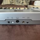 Detailní pohled na zadní panel stříbrného elektronického klávesového nástroje. Jsou zde vidět konektory a nápisy: --C+ 12V DC, USB s ikonou USB, MIDI…