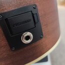 Detailní záběr na černý plastový panel s elektronikou osazený do dřevěného korpusu akustické kytary. Na panelu je vidět nápis FISHMAN a kovový výstup…