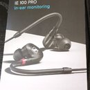 Fotografie znázorňuje krabici od sluchátek s nápisy SENNHEISER, IE 100 PRO a In-ear monitoring. Na obalu je zobrazen pár černých šupenek s ohýbatelný…