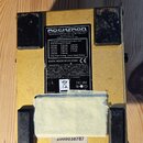 Zadní strana žlutého efekt pedálu značky ROCKTRON s nápisem TECHNOLOGY FOR GUITARISTS. Na spodní části je vidět bílý papírový štít překryté lepicí pá…