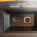 Detailní záběr na černý plastový panel s dvěma USB konektory umístěnými v prohlubni. Nad porty jsou bílé nápisy USB, MEMORY a COMPUTER doplněné o sym…