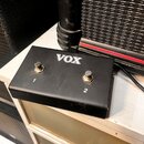 Černý kytarový pedál s nápisem VOX. Na horní straně jsou dvě stoupací tlačítka označená číslicemi 1 a 2. Povrch krabičky vykazuje drobné škrábance a…