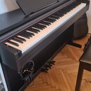Černý digitální piano s klávesnicí a integrovaným stojanem. Na krytu nad klávesami je bílý nápis KURZWEIL. Nástroj má tři zlaté pedály a k jeho boku…