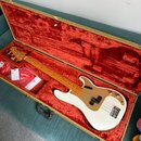 Bílá elektrická basová kytara v hnědém tvrdém kufru s červeným plyšovým vnitřkem. Na hmatníku je vidět nápis Fender a na krytce elektroniky jsou umís…