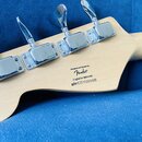 Záběr na hlavu basové kytary z přírodního světlého dřeva s chromovanými ladicími kolíky. Na zadní straně je černý nápis Designed and Backed by Fender…