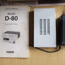Na fotografii je bílý manuál s nápisy Owner's Manual, Model D-80 a Digital Multitrack Recorder s logem fostex. Vedle manuálu je bílý kovový zdroj a č…