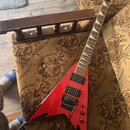 Červená elektrická kytara s tělem ve tvaru V a černými hranami. Na hlavě hmatníku je bílé logo Jackson. Kytara má dva černý humbuckery, černý most s…