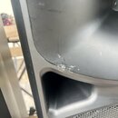 Detailní záběr na černý plastový povrch reproduktoru nebo ozvučňovacího systému. V místě ohybu materiálu nad výřezem je viditelné výrazné odřasání a…