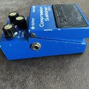 Modrý efekt pedál s nápisy Compression Sustainer a CS-2. Na horní části je černý spínač s nápisem BOSS a tři ovládací knoflíky, z nichž jeden je ozna…