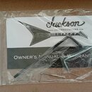 Průhledný plastový sáček obsahuje dokumentaci a nářadí. Na bílém papíře je černý nápis Jackson, adresa www.JACKSONGUITARS.com a silueta kytary s nápi…