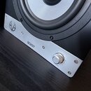 Detailní pohled na spodní část černého reproduktoru s přišroubovaným stříbrným kovovým panelem. Na panelu je vlevo logo ve tvaru stylizovaného trojúh…