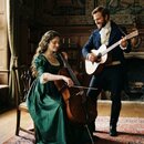 Na fotografii jsou dvě osoby v historických kostýmech s hudebními nástroji. Žena sedí na židli a hraje na dřevěné violoncello s hnědým povrchem. Muž…