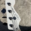 Fotografie zachycuje hlavu bílé basové kytary se čtyřmi černými ladicími kolíky. Na povrchu je černý nápis Squier BY FENDER a JAZZ BASS. Povrch je hl…